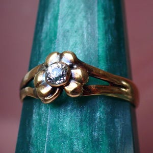 May include: A gold-toned ring with a flower-shaped design. The ring features a central, clear gemstone and four petal-like shapes around it. The ring is set against a green and pink background.