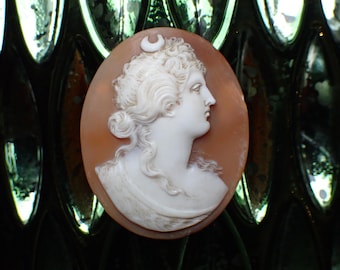 May include: Oval cameo pendant featuring a profile of a woman with curly hair and a crescent moon. The woman's image is carved in white, set against a light brown background. The cameo is set against a dark, textured background.