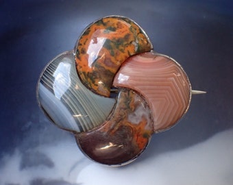 May include: A silver brooch featuring a four-leaf clover design crafted from agate stones. The stones are in shades of brown, orange, pink, and white.