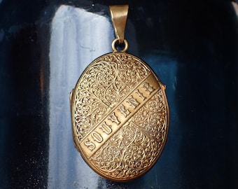 May include: A gold-toned oval locket with an intricate floral design and the word "SOUVENIR" engraved on the front.