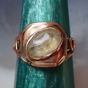 May include: A copper ring with a clear, faceted gemstone set in a bezel setting. The ring is on a green background.