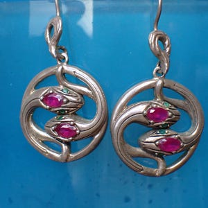 May include: Silver earrings with a distinctive design. Each earring has a circular frame with a stylized snake motif, incorporating two oval, ruby-coloured stones and small green accents. The earrings hang from a simple hook.