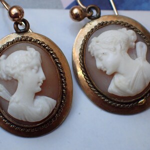 May include: A pair of gold tone earrings with cameo portraits of women. The cameos are set in oval frames with a rope-like border.