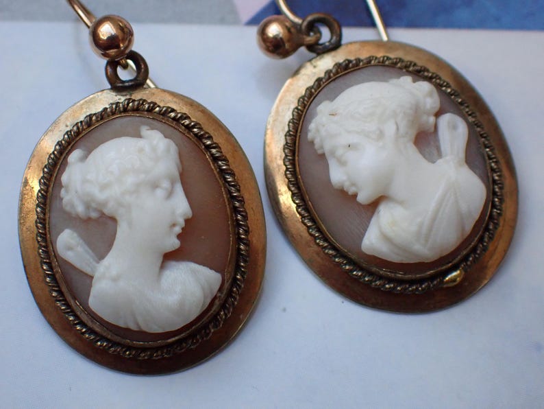 May include: A pair of gold tone earrings with cameo portraits of women. The cameos are set in oval frames with a rope-like border.