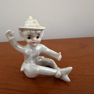 Op de afbeelding: Een wit keramieken beeldje van een zittende elf met een puntmuts. Het figuur heeft een parelmoerachtige afwerking, geschilderde gelaatstrekken waaronder donkere ogen en rode lippen, en is afgebeeld met één arm omhoog en de andere naar voren.