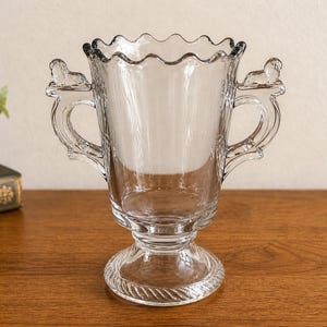 May include: Clear glass vase with a scalloped rim and two decorative handles. The vase has a pedestal base and is displayed on a wooden surface. The handles are adorned with small sculpted figures. A decorative piece for home decor.