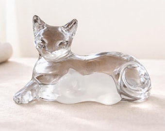 May include: A clear glass cat figurine, lying down with a smaller cat figure nestled beneath it. The sculpture is transparent, allowing light to pass through, and is set against a soft, neutral background.