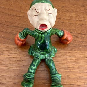 Pode incluir: Uma estatueta de cerâmica de um pequeno personagem semelhante a um elfo, vestindo uma roupa e gorro verdes com luvas de boxe laranja. A figura tem um rosto pálido com a boca bem aberta, lábios vermelhos e orelhas pontudas, sentada.