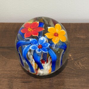 Peut inclure: Presse-papier en verre transparent avec trois fleurs color&eacute;es : rouge, jaune et bleue, avec des rubans bleus. Le presse-papier sph&eacute;rique repose sur une surface en bois.