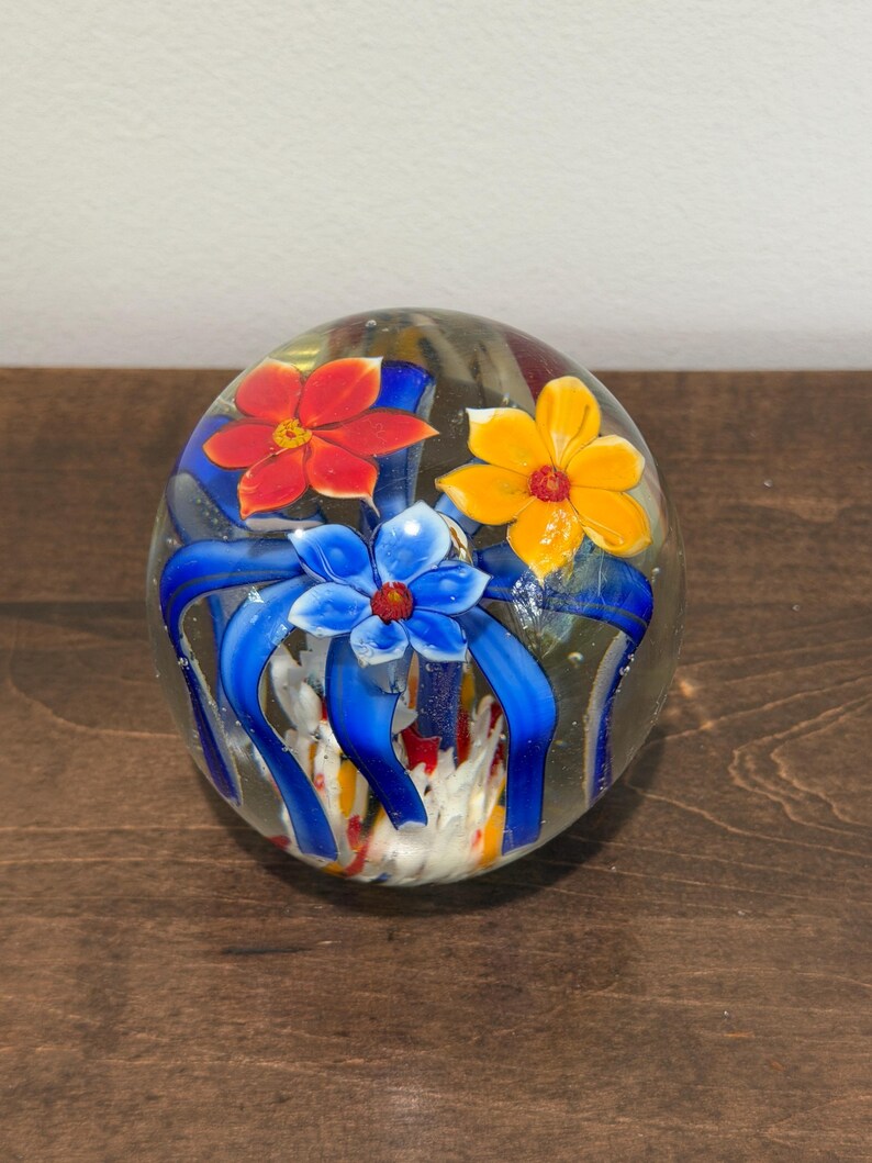 Peut inclure: Presse-papier en verre transparent avec trois fleurs color&eacute;es : rouge, jaune et bleue, avec des rubans bleus. Le presse-papier sph&eacute;rique repose sur une surface en bois.
