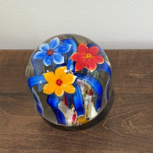 Peut inclure: Presse-papier en verre transparent avec trois fleurs color&eacute;es et des rubans bleus. Les fleurs sont bleues, rouges et jaunes. Le presse-papier est rond et repose sur une surface en bois fonc&eacute;. Environ 7.6 cm de diam&egrave;tre.