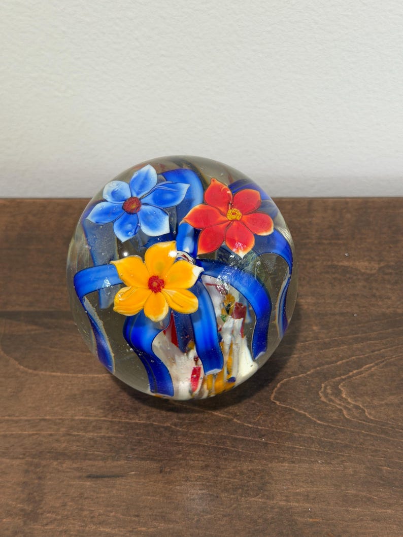 Peut inclure: Presse-papier en verre transparent avec trois fleurs color&eacute;es et des rubans bleus. Les fleurs sont bleues, rouges et jaunes. Le presse-papier est rond et repose sur une surface en bois fonc&eacute;. Environ 7.6 cm de diam&egrave;tre.