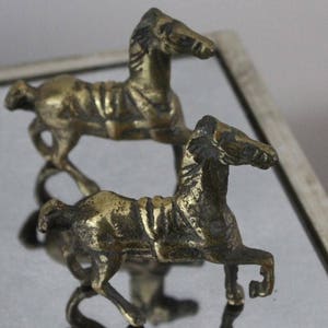 May include: Two small brass horse figurines in a trotting pose. Each metallic horse features a textured mane, a saddle detail on its back, and a polished finish that reflects light. The figures are positioned one behind the other.