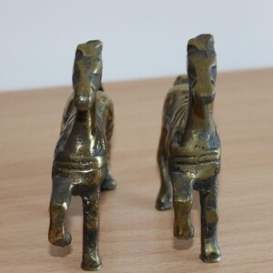 May include: A pair of brass horse figurines standing in a forward-facing position. Each metallic sculpture features a weathered, dark gold finish with visible texture and defined facial features, showcasing a standing stance with four legs.