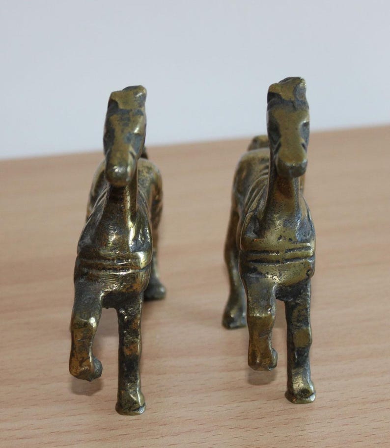 May include: A pair of brass horse figurines standing in a forward-facing position. Each metallic sculpture features a weathered, dark gold finish with visible texture and defined facial features, showcasing a standing stance with four legs.