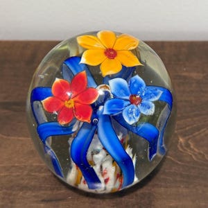 Peut inclure: Presse-papier en verre transparent avec trois fleurs color&eacute;es et des rubans bleus. Les fleurs sont rouges, jaunes et bleues, avec des centres d&eacute;taill&eacute;s. Objet d&eacute;coratif.