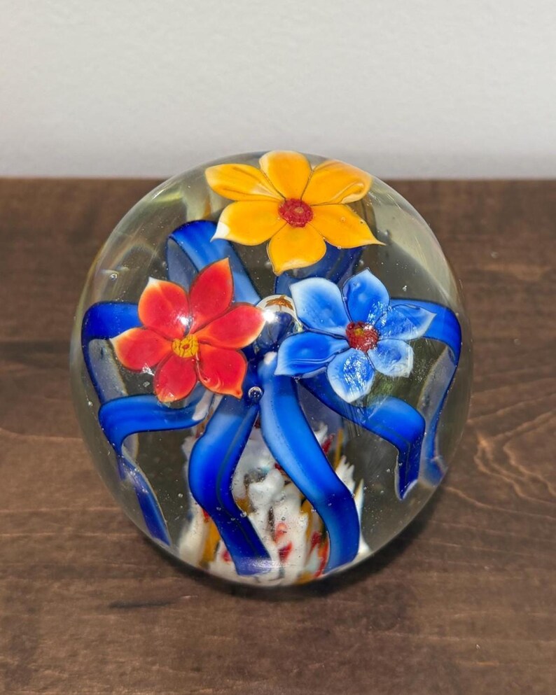 Peut inclure: Presse-papier en verre transparent avec trois fleurs color&eacute;es et des rubans bleus. Les fleurs sont rouges, jaunes et bleues, avec des centres d&eacute;taill&eacute;s. Objet d&eacute;coratif.