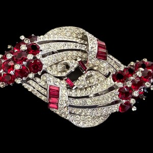 May include: A silver-toned brooch featuring a complex, swirling ribbon design encrusted with numerous clear, sparkling rhinestones. The piece is accented with clusters of deep red, round-cut stones and rectangular red baguette-cut stones.