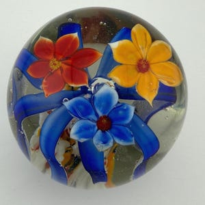 Peut inclure: Presse-papier en verre transparent avec trois fleurs color&eacute;es. Les fleurs sont rouges, jaunes et bleues, chacune avec un centre fonc&eacute;. Des rubans bleus entourent les fleurs. Le presse-papier est rond et l&eacute;g&egrave;rement bomb&eacute;.