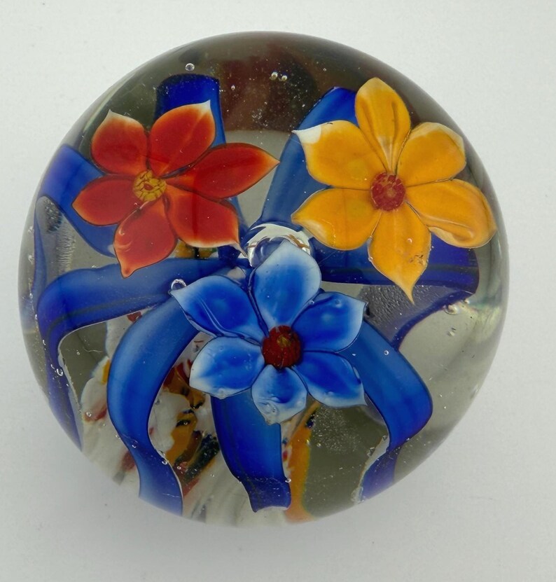 Peut inclure: Presse-papier en verre transparent avec trois fleurs color&eacute;es. Les fleurs sont rouges, jaunes et bleues, chacune avec un centre fonc&eacute;. Des rubans bleus entourent les fleurs. Le presse-papier est rond et l&eacute;g&egrave;rement bomb&eacute;.
