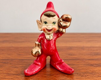 May include: A ceramic elf figurine wearing a bright red suit and matching pointed hat. The elf has a smiling face, pointed ears, and is wearing metallic gold boxing gloves on both hands. The figure is standing in a wide stance.