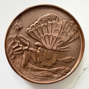 May include: A round, copper-colored decorative plate with a raised relief design. The design features a butterfly with detailed wing patterns, a flower, and foliage. The plate has a slightly textured surface and a warm, metallic hue.