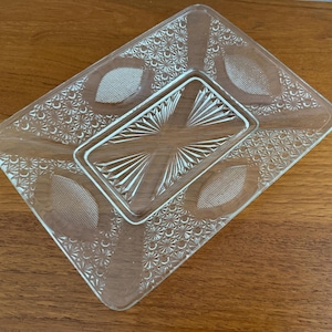 May include: Clear glass rectangular dish with a raised rectangular center. The dish features a detailed pattern of small flowers and oval shapes. The center has a starburst design. The dish is on a wooden surface.