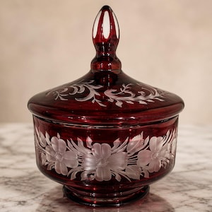 May include: A ruby red glass jar with a lid and a decorative floral design. The jar features etched white flowers around the base and on the lid. The finial is white with red accents.