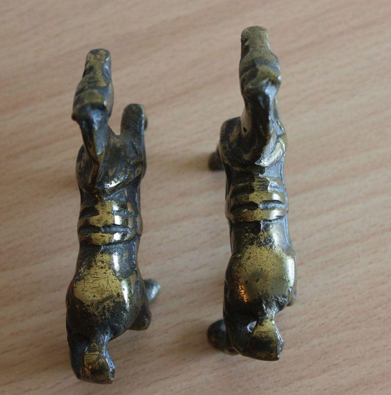 May include: Two small, weathered brass horse figurines shown from a top-down perspective. Each metallic horse is depicted in a rearing position with visible textured details along the back and mane, showcasing a dark, aged patina finish.