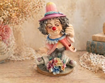 May include: A whimsical clown figurine with a pink hat and a red nose. The figurine is wearing a pink and blue outfit and is seated on a base adorned with colorful flowers. The clown has curly gray hair and a smiling expression.