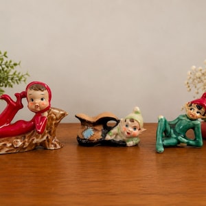 Puede incluir: Tres figuras de cerámica con personajes caprichosos. Una figura lleva una capucha roja y yace sobre un tronco marrón. La figura central es un pequeño elfo descansando dentro de un zapato marrón. La tercera figura es un elfo verde recostado.
