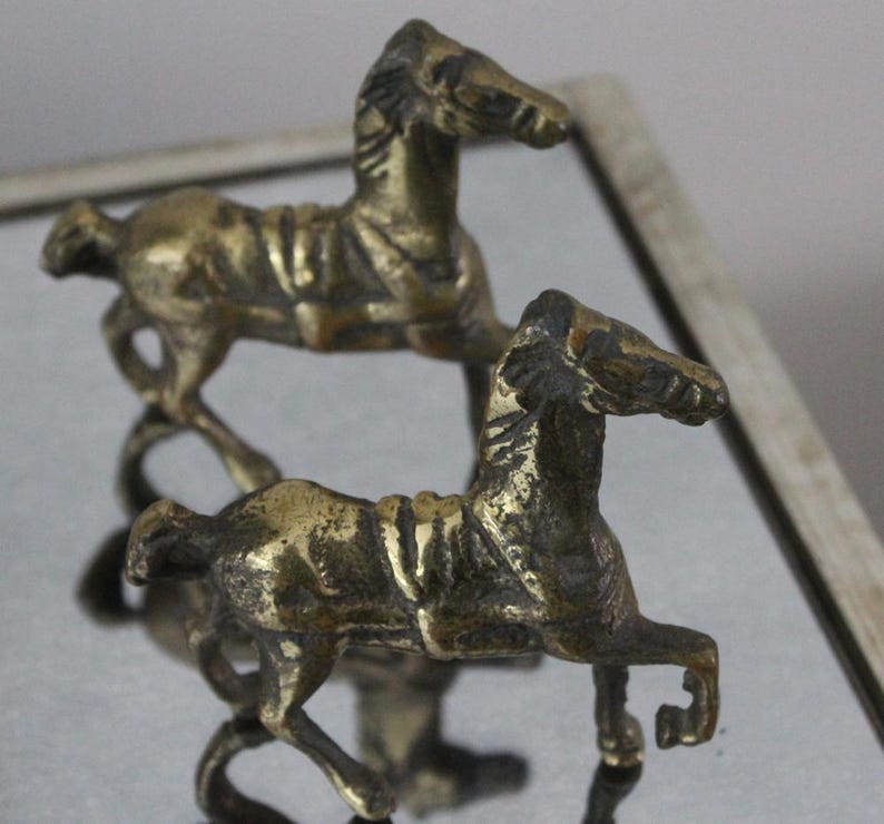 May include: Two small, metallic, brass-colored horse figurines standing in a side-profile pose. Each horse features a textured mane, a saddle detail on its back, and a raised front leg, showcasing a weathered, aged patina finish.