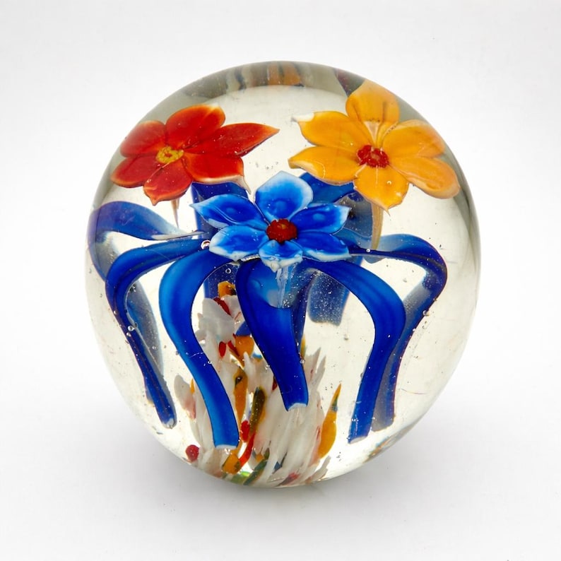 Peut inclure: Un presse-papier en verre transparent avec des motifs floraux color&eacute;s. Le presse-papier pr&eacute;sente trois fleurs rouges, jaunes et bleues, avec des accents bleus en forme de ruban. Les fleurs ont des centres rouges et sont enferm&eacute;es dans une sph&egrave;re de verre transparente.