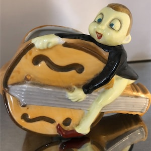 Peut inclure: Une figurine en céramique d'une personne aux grands yeux et un large sourire, tenant un instrument de musique jaune et brun. La figurine est un pot de fleurs avec un trou en haut pour planter.