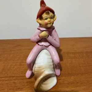Op de afbeelding: Een keramieken beeldje van een elf met een rode puntmuts en een lichtroze outfit met donkerrode randen. De elf zit bovenop een witte en beige schelp. Het figuurtje heeft geschilderde gelaatstrekken, waaronder roze wangen en bruin haar.