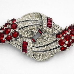 Könnte beinhalten: Eine silberfarbene Vintage-Brosche mit einem auffälligen Design. Die Brosche ist mit klaren Kristallen, roten rubinartigen Steinen und rechteckigen roten Akzenten besetzt. An jedem Ende befinden sich rote Steingruppen.