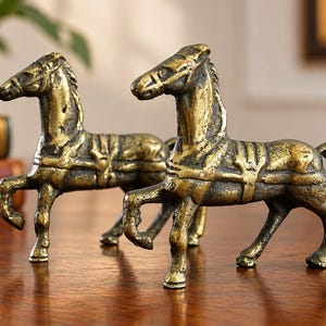 May include: A pair of brass horse figurines, each depicted in a standing pose with a harness and saddle detail. The metal has a weathered, textured finish with dark patina accents in the crevices, showcasing a metallic gold-toned surface.
