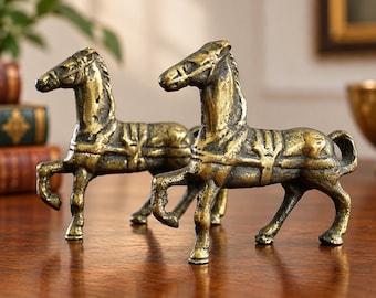 May include: A pair of brass horse figurines, each depicted in a standing pose with a harness and saddle detail. The metal has a weathered, textured finish with dark patina accents in the crevices, showcasing a metallic gold-toned surface.