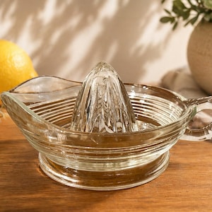 Op de afbeelding: Een heldere glazen citruspers met een ruimer en een schenktuit. Twee gele citroenen zijn zichtbaar op de achtergrond. De pers staat op een houten oppervlak, met een decoratieve plant en een vaas op de achtergrond.