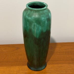May include: A tall, cylindrical ceramic vase featuring a smooth, rounded rim and a tapered base. The exterior surface displays a blended gradient of deep forest green at the bottom, transitioning into a lighter, vibrant emerald green toward the top.