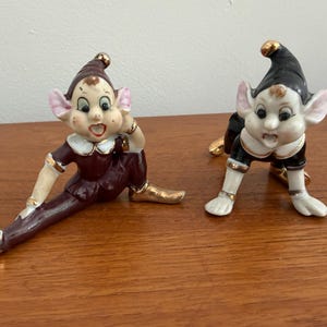May include: Two ceramic elf figurines with painted facial features, large ears, and gold-trimmed clothing. One elf wears a dark maroon outfit and sits with legs extended, while the other wears a black outfit and is positioned in a crawling pose.