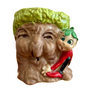 May include: A vintage ceramic planter shaped like a tree with a face, featuring a green leafy top and a small elf figure in red and black clothing. The planter has a detailed, textured surface.