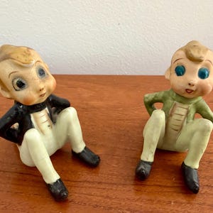 May include: Two ceramic figurines of seated characters with large blue eyes and light brown hair. One figure wears a black jacket with white trousers, while the other wears a green jacket with light green trousers. Both have painted facial features.