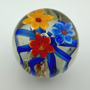 Peut inclure: Presse-papier en verre transparent avec trois fleurs color&eacute;es et des rubans bleus. Les fleurs sont jaunes, rouges et bleues, avec des p&eacute;tales et des centres d&eacute;taill&eacute;s. Le presse-papier est rond et d&eacute;coratif.