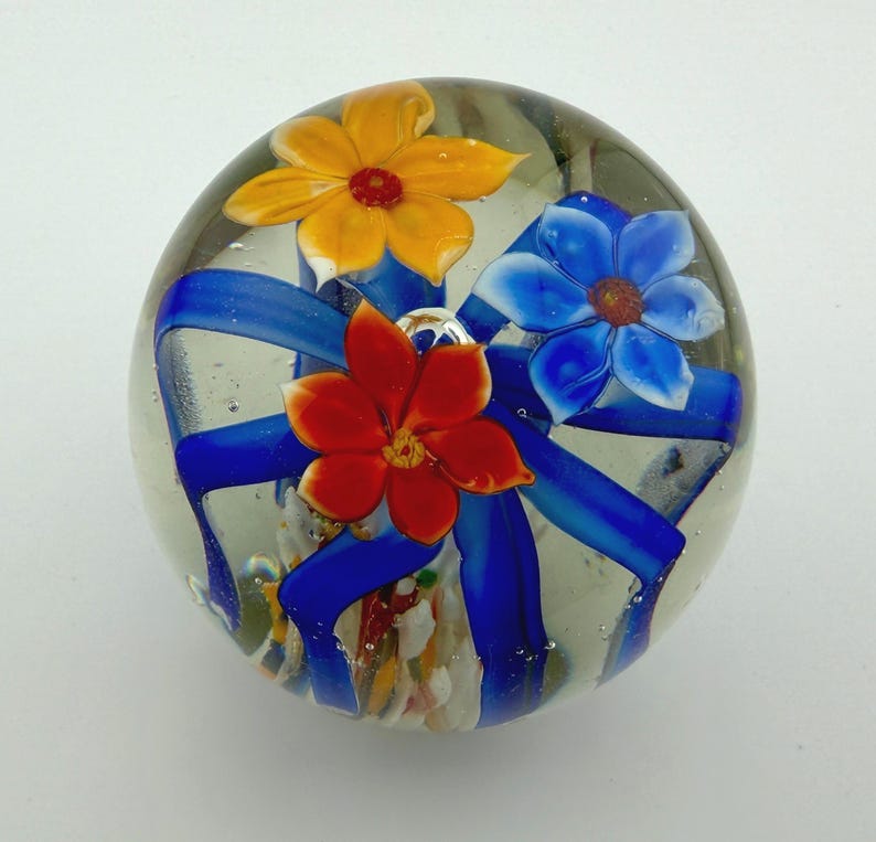 Peut inclure: Presse-papier en verre transparent avec trois fleurs color&eacute;es et des rubans bleus. Les fleurs sont jaunes, rouges et bleues, avec des p&eacute;tales et des centres d&eacute;taill&eacute;s. Le presse-papier est rond et d&eacute;coratif.