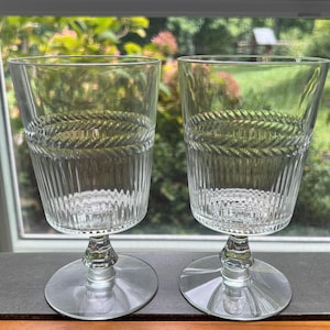 Puede incluir: Dos copas de vidrio transparente con un patrón grabado decorativo. Las copas tienen una copa alta y cilíndrica y un tallo corto.