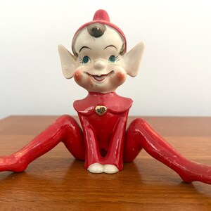 May include: A ceramic elf figurine with a glossy red outfit, pointed hat, and curled shoes. The elf has a pale face, large pointed ears, rosy cheeks, and a small gold heart emblem on the collar. It is posed in a seated position with legs extended.