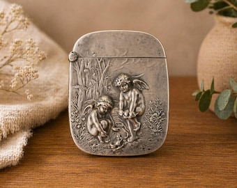 May include: A silver-toned match safe with a hinged lid. The front features a detailed relief of two cherubs in a garden setting. The cherubs are depicted with wings and are interacting with each other. The background includes trees and foliage.
