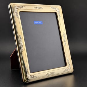 May include: A rectangular gold-toned picture frame with ornate engraved scrollwork patterns at each corner and along the edges. The frame features a dark interior display area with a small blue rectangular label reading "Argento 925".
