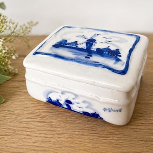 May include: A white ceramic trinket box with a blue and white windmill design on the lid. The box has a rectangular shape with rounded edges and a matching blue border. The sides of the box also feature blue painted details.
